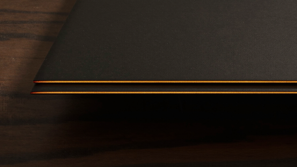 Extreme macro of two razor-thin matte black device edges stacked on dark walnut, each catching a warm amber specular line, illustrating the thickness difference between a folded and unfolded foldable phone